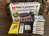 Atari Flashback 7 console box, yellow Superman game box, white iClicker 2 remote, and stack of Nintendo DS game cases, placed on a metal table.
