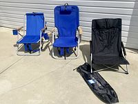 Front view of three folding beach chairs on concrete. Two blue chairs with wooden armrests and one black chair. LA Kings logo storage bag is next to black chair.