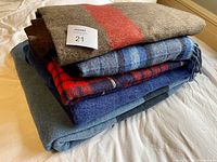Stack of five folded blankets showing various colors including brown with red stripe, multiple shades of blue with different patterns and textures