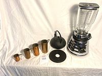 Full lot display showing all items: vintage Osterizer blender, Japanese cast iron teapot with trivet, and five stacking copper/brass measuring cups.