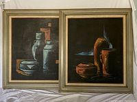 Pair of framed canvas paintings side by side resting on white fabric backdrop, showing compositions of pottery forms on dark background.