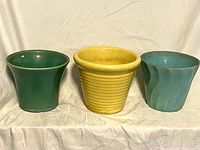 Three ceramic flower pots in green, yellow, and blue placed side by side on white cloth background.