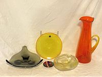 Group photo showing all six colored art glass pieces together: orange pitcher, yellow dish, dark smoked dish, greenish dish, and two smaller glass items.