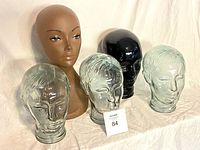 Group photo of five clear glass display heads, one black glass display head, and one painted plastic display head on a white cloth background.