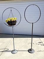 Two vintage brass garden hoops on stands, one with a hanging basket attached, shown side by side outdoors on concrete.