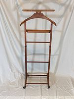 Full view of the wooden valet stand showing hanger top, trouser bar, additional horizontal bars, and caster wheels at base.