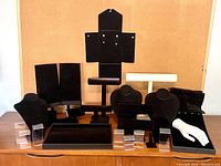 Photo of black velvet necklace busts, bracelet bars, ring stands, clear lucite earring holders, and a white hand-shaped jewelry display arranged on a wooden surface.