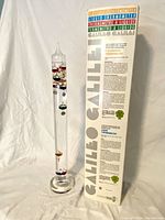 Full view of the large Galileo liquid thermometer with original box standing vertically. The glass tube has multiple colored bulbs and a round base.