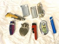 Nine assorted lighters displayed on white fabric background showing different shapes, styles and materials including novelty and classic designs.