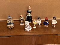 Eight assorted porcelain and ceramic figurines displayed on wood surface against neutral background showing variety of designs and styles.