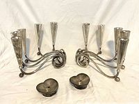 Front angled view of two silver tone triple arm candle holders and two small heart-shaped candle trays on white backdrop.
