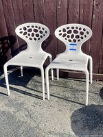 Two lightweight off-white plastic chairs with oval perforated backrests, sitting on gravel outdoors against a wooden fence