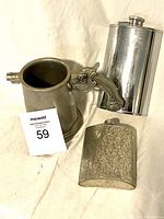 Pewter beer stein with pistol-style handle beside two metal flasks, one polished and one embossed with floral pattern.