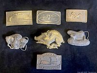 Top side front view of nine solid brass belt buckles arranged on a black background showing detailed embossing and figure shapes