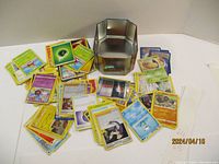 Photo showing a large assortment of Pokemon trading cards spread around a hexagonal metal tin storage box. Cards include multiple types and some with holographic foil.