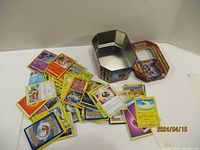 Photo showing over 110 assorted Pokemon trading cards spread out next to a Pokemon-themed metal tin with removable lid.