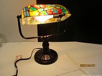 Lit stained glass banker’s lamp showing colorful hummingbird and flower design on shade with bronze-tone base.