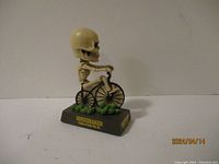 Side view of bobble head skeleton riding a bicycle on labeled base