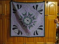 Full view of the square quilt hanging against a wooden background. The quilt features a central floral applique design with black fan shapes in each corner and green leaf shapes around the center flower.