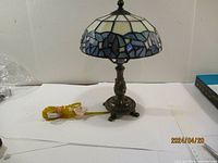 Side and angle view of small Tiffany style lamp showing stained glass shade with white, blue, and purple glass pieces and ornate metal base with three feet and yellow cord.