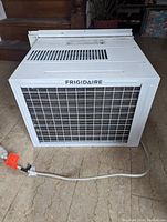 Frigidaire air conditioner unit front facing with grille and power cord visible on floor.