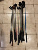 Top view of 12 assorted golf clubs with visible shafts and grips laid side-by-side on floor tile.