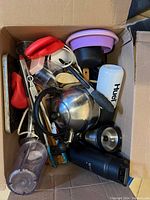 Open box showing assorted kitchen items including a stainless steel teapot, a white Huel cup, kitchen utensils, silicone lids, and other small kitchen tools.