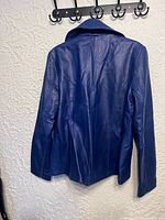 Back view of the women's blue genuine leather jacket hanging on hooks, showing the jacket's smooth leather texture and tailoring details.
