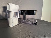 Clear plastic Brita filter pitcher with white lid containing two extra white filter cartridges inside, and next to it clear Brita in-fridge water dispenser with black lid and silver spigot