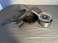 Set of one cast iron pan, one small stainless steel pot with glass lid, and one non-stick large frying pan on a dark surface.