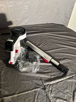 White and black cordless vacuum with clear dust container and metal extension wand, plastic accessories wrapped in plastic on a grey surface.