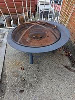 Top angled view of the rusty metal fire pit showing mesh screen top and three legs on concrete surface with a dog behind a metal gate in background.