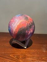 Front view of the spherical galaxy lamp showing bright red and purple nebula and star patterns on textured surface resting on a white angular stand.