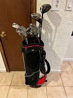 Full view of the golf bag with clubs inside standing on tile floor against wooden door and white textured wall.
