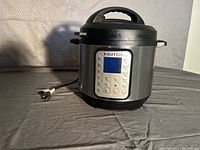 Front view of Instant Pot showing digital control panel, lid, and handles, power cord visible.