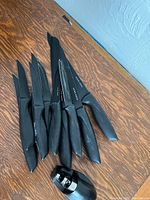 Six black-handled kitchen and steak knives laid out on a wooden surface along with a black handheld knife sharpener.