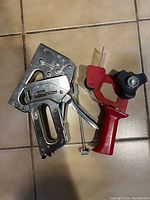 Two staplers (a metal Stanley Heavy Duty Tacker and a chrome hand stapler) next to a red-handled Scotch tape dispenser on a tiled floor.