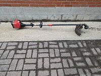 Photo showing the full side view of the Troy-Bilt weed wacker placed horizontally, highlighting the red engine and straight shaft with handle grip.
