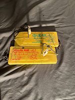 Two yellow plastic Yellow Bird fishing accessory cases with text and images on top, attached cords, clips and toggles visible.