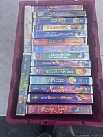 Photo showing 16 VHS tapes in pink plastic bin, with visible titles including Pocahontas, Beauty and the Beast, Aladdin, Lion King II, Snow White, Winnie the Pooh, Return of Jafar, and others.