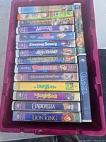 Photo of 16 VHS tapes stacked in a red plastic bin, showing titles including Walt Disney classics like 101 Dalmatians, Sleeping Beauty, Lady and the Tramp, The Lion King, and others.