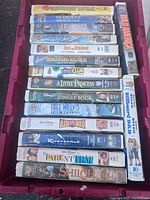 Photo showing 16 VHS tape cases stacked in one box with visible titles including Stuart Little, Free Willy 2 and 3, Air Bud, Jungle Book and more.
