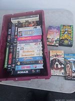 Photo showing 25 VHS tapes in a red plastic bin and some stacked beside it on a surface. Tapes are identifiable by visible cover art and titles.