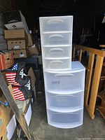 Two plastic storage containers side by side, showing front facing drawers and handles, in a storage space with boxes and other items around.