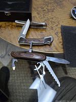 Three collectible stainless steel pocket knives shown on a wood table, two single blade knives and one multi-tool knife with various implements extended