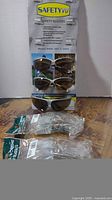 Photo showing 3 pairs of SafetyVu dark tinted safety glasses on a backing card and 2 pairs of clear industrial safety glasses in packaging on a wood surface.