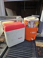 View of two coolers, an orange Igloo 5-gallon beverage cooler with a white lid and attached gray cup dispenser, and a white and red Gott Tote 18 cooler. Both appear to be lightly used.