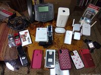 Full table view showing all items included in the lot placed on a wooden surface: vintage flip phones, Apple networking tower, Apple mouse, Netgear devices, phone cases, IP phone, and cables.