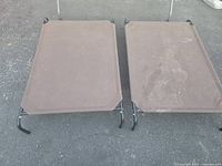 Two elevated pet beds placed on asphalt. Fabric surfaces are brown and show signs of dirt and use. Frames are black metal tubular with four legs each.