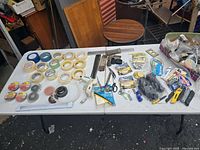 Wide top-down photo showing the full set of assorted tapes, tools, hardware, and office repair items laid out on a white table outdoors.